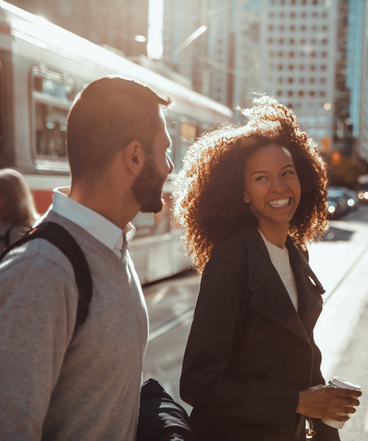 Un homme et une femme se regardent en souriant. La femme tient une tasse de café et l'homme porte un sac à dos. À l'arrière-plan, des personnes marchent vers un tramway au centre-ville de Toronto.