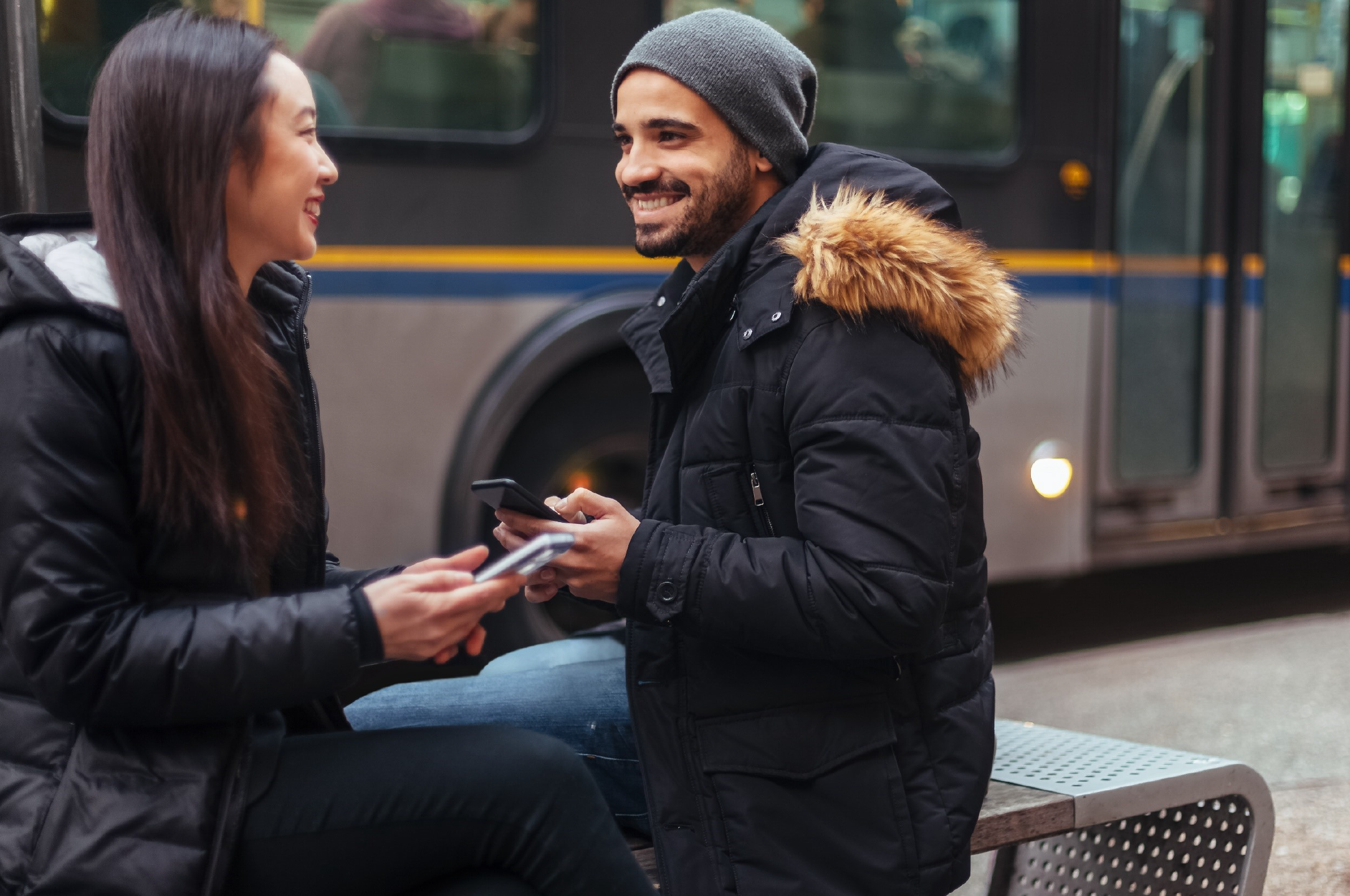 Deux personnes se souriant l'une à l'autre, assises sur un banc et tenant leurs téléphones, avec un autobus passant derrière elles.