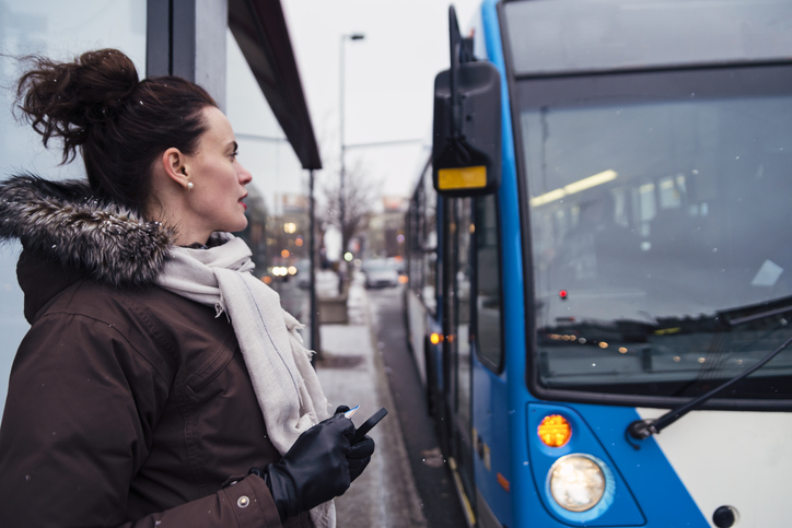 Une femme tient son téléphone et sa carte de transport alors qu’un autobus s’arrête. La neige tombe légèrement.