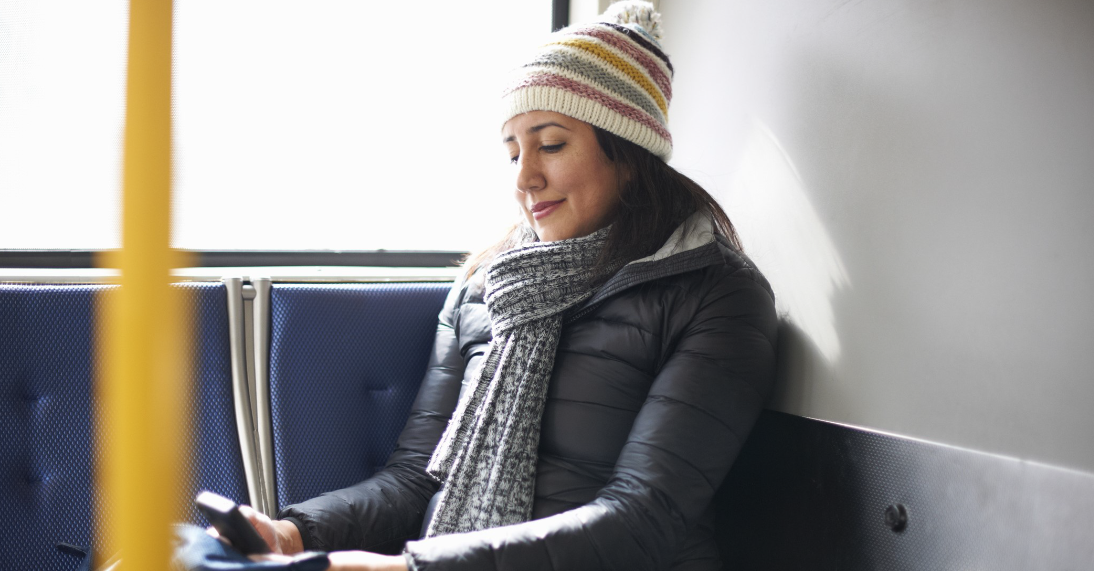 Une femme portant un chapeau et une écharpe d'hiver sourit à son téléphone pendant qu'elle est assise dans un autobus.