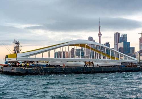 Un chaland transporte un pont moderne sur l’eau. On voit l’horizon de la ville de Toronto en arrière-plan.