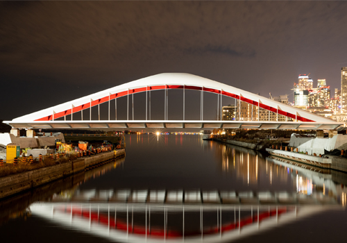 Un pont moderne illuminé dans la nuit. On voit la ligne d’horizon de la ville en arrière-plan.