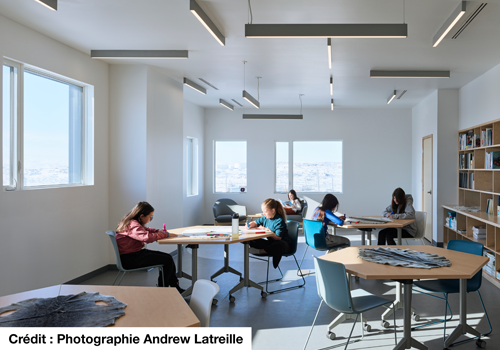 Des enfants étudient, installés à des tables rondes dans une pièce lumineuse. Une grande étagère remplie de livres est adossée à un mur.