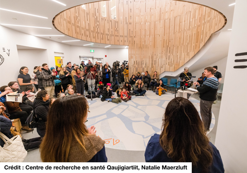 Un rassemblement communautaire dans une grande salle circulaire lambrissée de bois. Un orateur devant un lutrin s’adresse à l’auditoire.