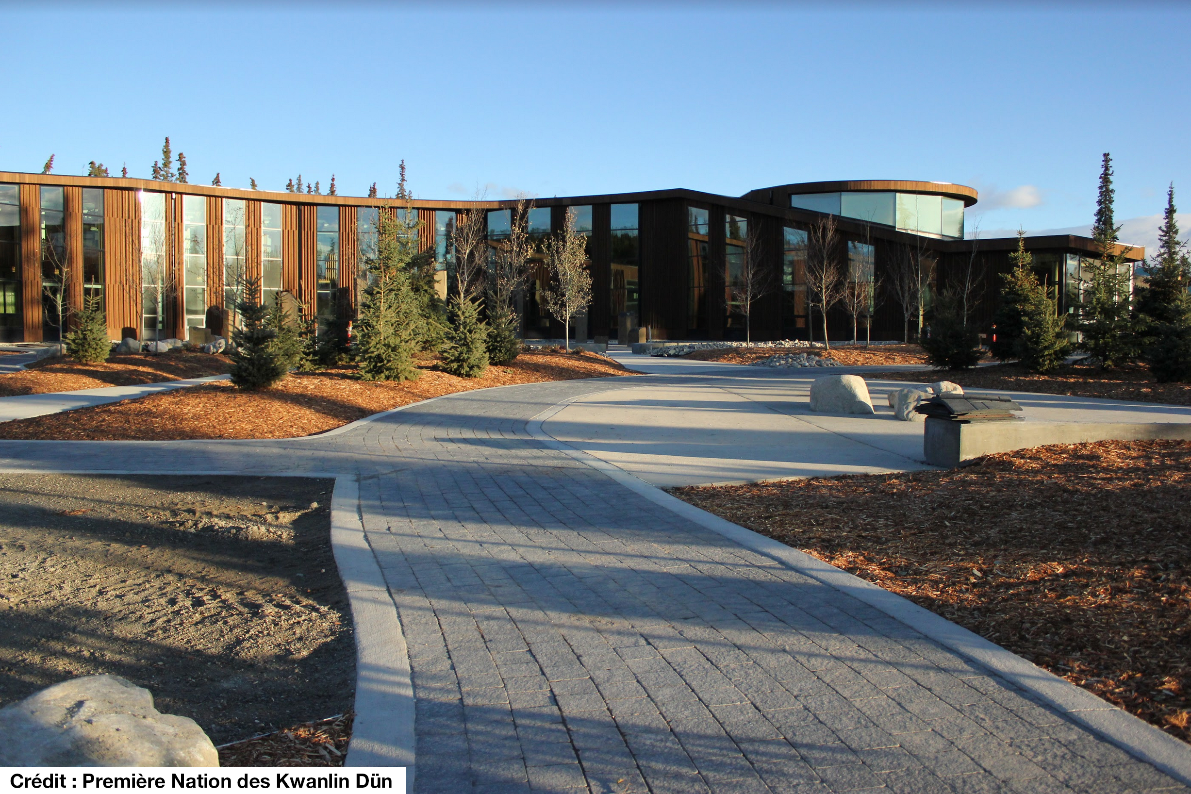 Grand bâtiment communautaire en bois avec des fenêtres du sol au plafond. Allée pavée et aménagement paysager devant le bâtiment. 