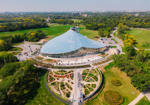 Vue aérienne d’un grand bâtiment au toit de verre, entouré de jardins et d’espaces verts.